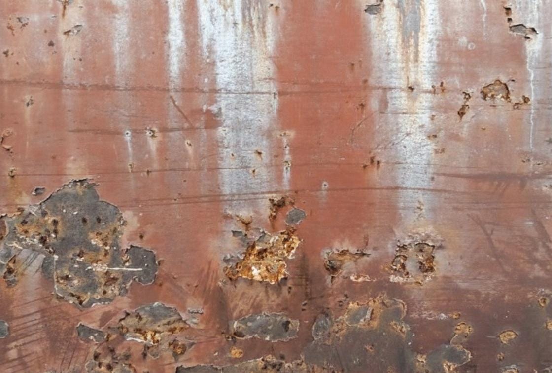 Macro de chapa metálica com corrosão severa por pites e descamação, ilustrando falhas em aços inoxidáveis comuns.