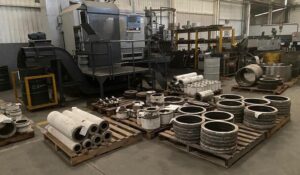 Interior industrial com peças fundidas organizadas em pallets e máquinas ao fundo, mostrando a atuação da TCINOX em fundição de ligas especiais com foco em qualidade e precisão.