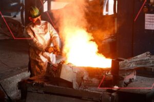 Operador da TCINOX com traje aluminizado de proteção manuseia metal incandescente em forno industrial, com fumaça e brilho intenso de calor.